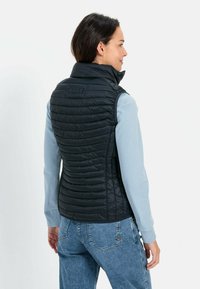 Zwarte gewatteerde vest met een hoge kraag, met een gestikte textuur en een getailleerd ontwerp, gedragen over een lichtblauw langemouwen shirt.