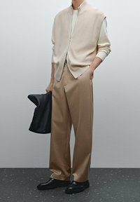 Beige Weste mit Reißverschluss über einem cremefarbenen Oberteil, kombiniert mit hellbraunen Hosen mit weitem Bein und schwarzen Schuhen. Hält eine schwarze, strukturierte Ledertasche.