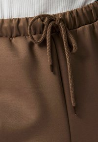 Pruunid elastse vöökohaga püksid, millel on seotud pael ja nähtav kangastruktuur, kombineeritud valge soonikkoega topiga.