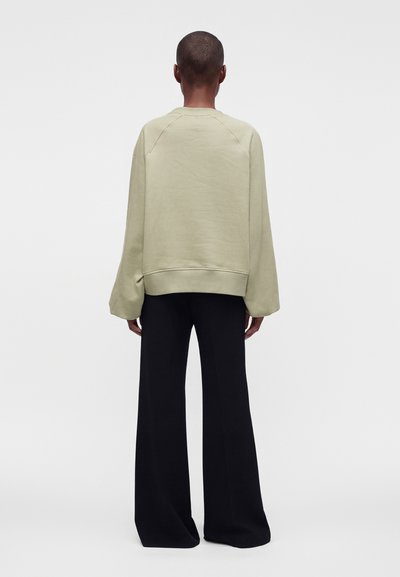 Personne de dos, portant un sweatshirt vert clair ample et un pantalon noir à jambes larges, debout devant un fond clair et uni.