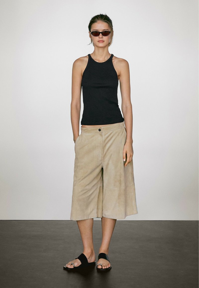 Top senza maniche nero, pantaloni morbidi beige a gamba larga e sandali neri. Modella in piedi contro uno sfondo neutro. Tessuto morbido con vestibilità ampia.
