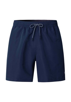 Hombre sin camiseta que lleva pantalones cortos de baño azul marino y zapatos beige sin cordones, de pie contra un fondo liso color gris claro.