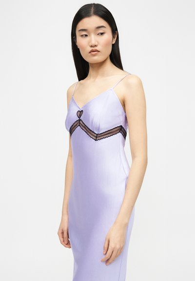 Νεαρή γυναίκα με λιλά φόρεμα-λάσπη με λεπτά τιράντες και μαύρη δαντελένια καρδιά, στέκεται μπροστά σε απλό φωτεινό φόντο.