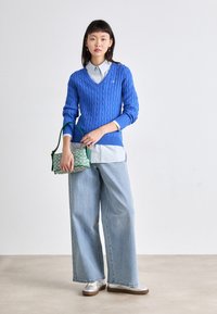 Suéter azul de punto con cuello en V, superpuesto a una camisa de collar azul claro, combinado con jeans de pierna ancha azul claro y un bolso verde con estampado.