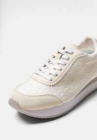 Zapatilla de color beige claro con parte superior texturizada que presenta un patrón floral, cordones planos y una suela de goma blanca. Punta redonda y interior acolchado.