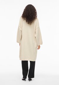 Γυναίκα με σγουρά μαλλιά που φοράει ένα μακρύ μπεζ παλτό πάνω από μαύρο παντελόνι και μαύρες γόβες, στέκεται με την πλάτη γυρισμένη σε λευκό φόντο.