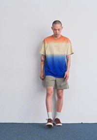 Nike gradient marškinėliai, pereinantys iš oranžinės į mėlyną, derinami su smėlio spalvos šortais su logotipo atspaudu. Dėvimi su baltais kojiniais ir rudos spalvos batais.