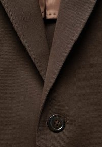 Brauner Stoffblazer mit Reverskragen, sichtbaren Nähten und einem runden, dunklen Knopf mit vier Löchern. Glatte Textur.