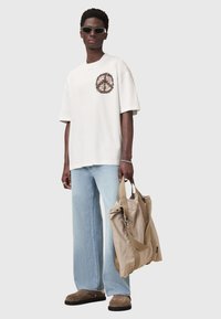 Hvid oversized t-shirt med et cirkulært grafisk design på brystet, parret med lyseblå bredbenede jeans og en beige taske. Brune slip-on sko.