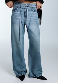 Světle modré džíny s širokými nohavicemi z denimu, s vázáním v pase a předními kapsami. Dokončené černými botami se špičatou špičkou.