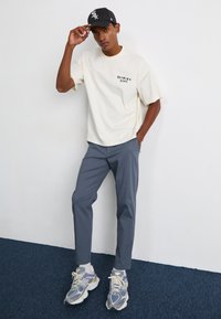 Cream short-sleeve t-shirt with a logo on the left chest, paired with slim dark gray pants and gray-silver sneakers with purple accents.