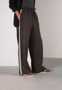 Weit geschnittene Hosen aus dunkelbraunem Stoff mit weicher Textur, ausgestattet mit weißen Seitenstreifen und einem elastischen Bund mit Kordelzug.