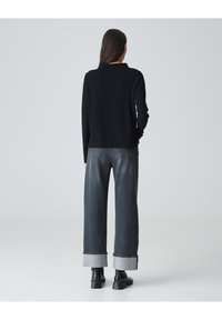 Schwarzer strukturierter Pullover mit langen Ärmeln, kombiniert mit grauen weit geschnittenen Jeans, die umgeschlagene Säume haben, und schwarzen Knöchelschuhen.