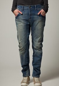 Modré džíny z denimu s vybledlým povrchem, rovný střih, pět kapes a zapínání na knoflík. Nosené se světlými teniskami.