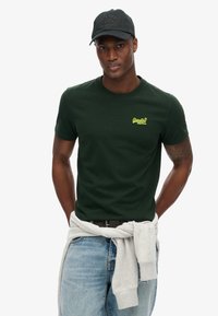 Green cotton t-shirt with a round neckline and yellow logo details, paired with light blue denim jeans and a grey sweatshirt tied at the waist.