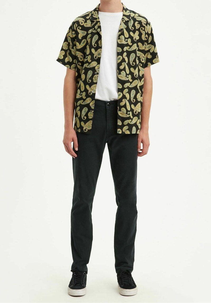 Camicia nera a maniche corte con motivo paisley giallo, abbinata a t-shirt bianca e pantaloni aderenti neri, completata da sneakers nere.