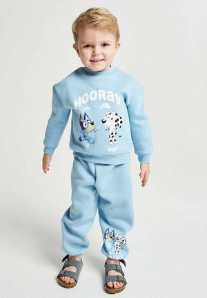 Young boy smiling, wearing light blue sweatpants and sweatshirt with "Hooray" and cartoon dog prints, standing on plain white background.