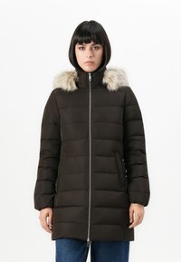 Kvinde med kort sort hår iført en sort quiltet vinterjakke med pelsbesat hætte og blå jeans, stående foran en hvid baggrund.