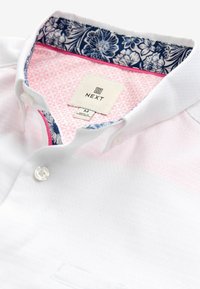 Polo shirt bianco con un motivo testurizzato, dettagli rosa all'interno del colletto e una stampa floreale blu navy nella parte interna del collo. Etichetta visibile.