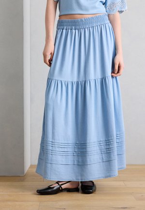 Kobieta nosząca jasnoniebieską, warstwową maxi spódnicę z plisami przy krawędzi oraz czarne baleriny Mary Jane na drewnianej podłodze.