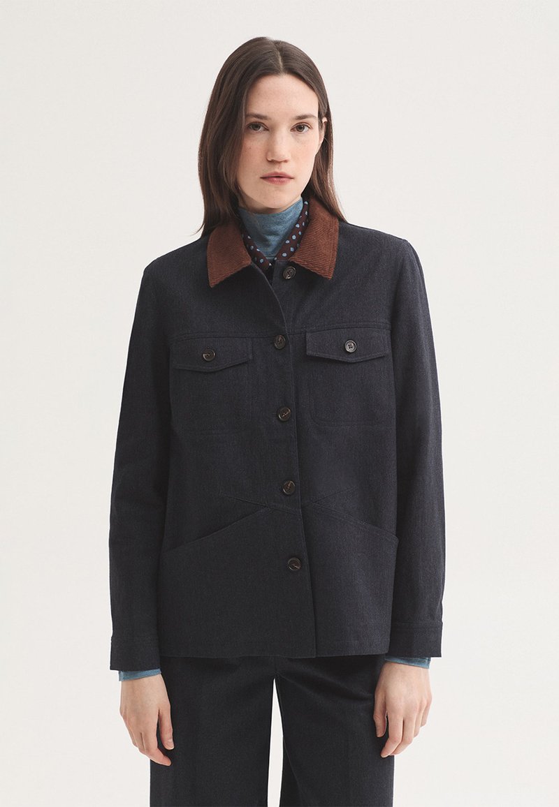 Femme portant une veste sombre boutonnée avec un col en velours côtelé marron, superposée à un col roulé bleu et un pantalon noir, debout sur un fond blanc.