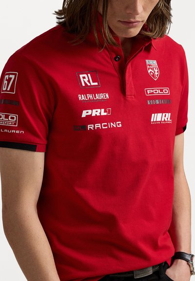Červený polo tričko s límečkem, zdobené několika logy v černobílé. Krátké rukávy s kontrastními černými akcenty. Hladká textura.