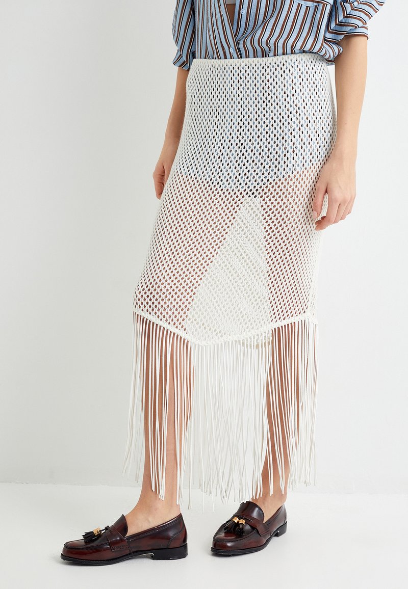 Jupe en maille blanche avec un ourlet à franges, présentant un design en tricot ouvert. Portée avec des mocassins marron, mettant en avant un tissu texturé et un motif horizontal.