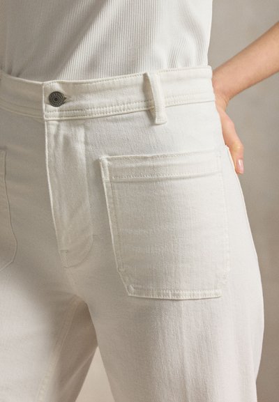 Κοντινό πλάνο ενός ατόμου που φοράει λευκά τζιν με ψηλή μέση και μεταλλικό κουμπί, δείχνοντας μπροστινή τσέπη και χέρι που ακουμπάει στο ισχίο.