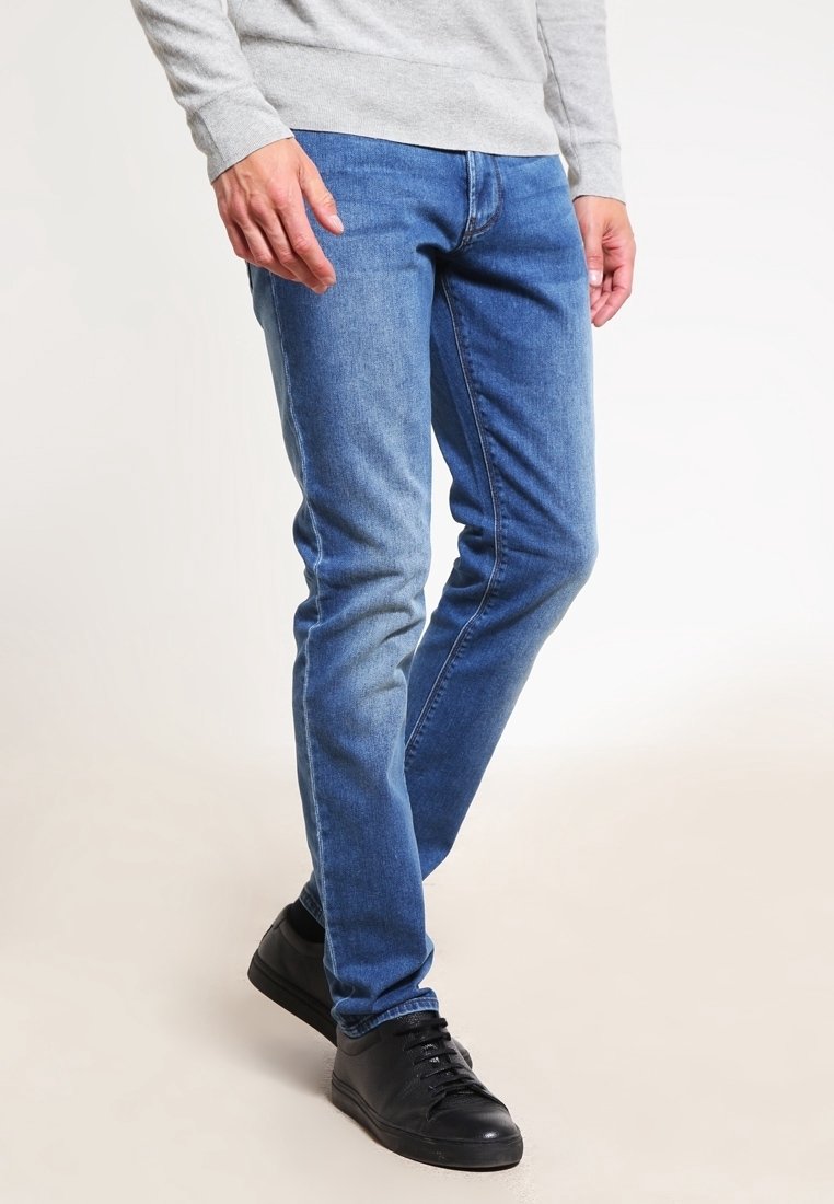 Faded blue denim jeans with a slim fit, featuring five pockets and a zip fly. Worn with a gray long-sleeve top and black sneakers.