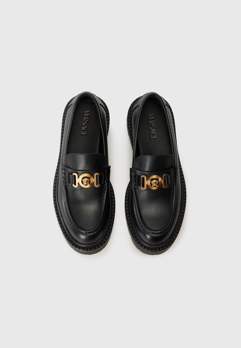 Mocassins en cuir noir avec un accent en chaîne dorée, bout rond et semelle texturée, conçus avec un profil bas et une finition lisse.