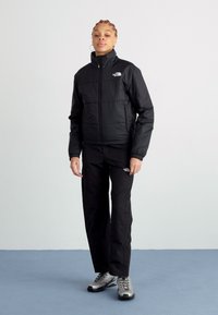 Giacca nera isolante con colletto alto, zip frontale e logo, abbinata a pantaloni neri coordinati e scarpe da hiking argentate.