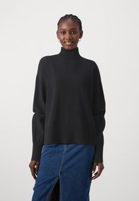 Pull noir à col roulé avec des manches larges et ourlet côtelé, associé à une jupe en denim avec une fente frontale, sur un fond neutre.