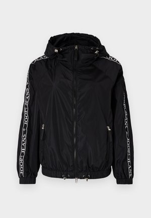 Zwarte nylon jas met een rits aan de voorkant, capuchon, elastische manchetten en een trekkoord onderaan. Voorzien van witte merkstrepen langs de mouwen.