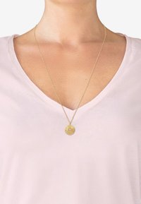 Gylden vedhæng halskæde på en tynd kæde, med indviklede cirkulære mønstre, båret over en lys pink v-hals t-shirt.