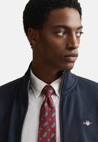 Marineblauwe jas met rits over een wit overhemd, gecombineerd met een bordeauxrode stropdas met blauwe paisley patronen. GANT-logo op de borst van de jas.