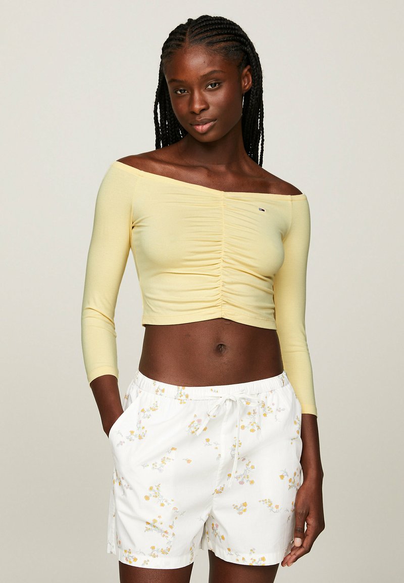 Helle gelbe schulterfreie Crop-Top mit gerafften Details, kombiniert mit weißen Shorts mit einem Blumenmuster und einem Kordelzugbund.