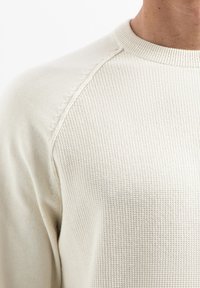 Licht crèmekleurige gebreide trui met een gestructureerd patroon, ronde halslijn en raglanmouwen met zichtbare stikdetails.