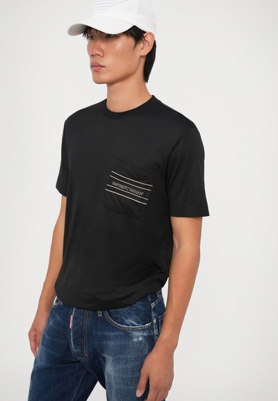 T-shirt preta de manga curta com um bolso, apresentando detalhes em riscas brancas. Combinada com jeans de ganga azuis com um acabamento desgastado.