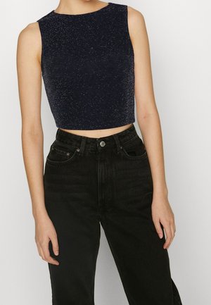 Kobieta ubrana w bezrękawnikowy, ciemnoniebieski krótki top i czarne, wysokie jeansy, stojąca na tle jasnego, jednolitego tła.