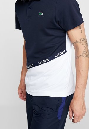 Uomo che indossa una polo Lacoste blu navy e bianca con logo e pantaloncini neri, mostrando il braccio destro tatuato su sfondo uniforme.