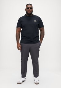Tricou din bumbac negru cu logo pe față, asortat cu pantaloni cargo gri cu buzunare laterale și adidași albi cu accente negre și maronii.