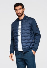 Mand med mørkt hår og skæg iført en marineblå quiltet jakke over en hvid t-shirt med print og blå jeans, stående foran en ensfarvet baggrund.