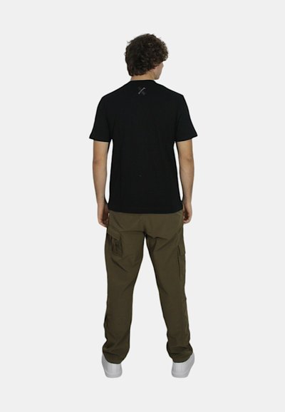 T-shirt nera a maniche corte con scollatura rotonda. Pantaloni cargo verde oliva con molte tasche. Sneakers bianche. Sfondo semplice.