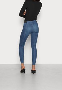 Persona con jeans ajustados bicolor azul, camiseta negra de manga larga y tacones negros de espalda descubierta, de pie con los brazos cruzados y la espalda hacia la cámara.