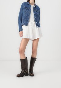 Chaqueta de mezclilla en azul oscuro con botones plateados, combinada con un vestido corto blanco y botas de cuero marrón hasta la rodilla con detalles de hebilla.