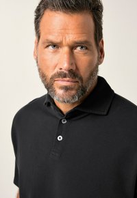Černé polo tričko vyrobené z texturované tkaniny, s límcem a tříknoflíkovou légou se tmavými knoflíky. Detailní záběr na horní část trička.
