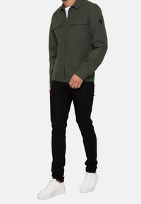 Veste zippée couleur vert olive avec deux poches poitrine, un col et une finition en tissu lisse, associée à un jean noir slim et des baskets blanches.