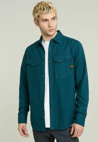 Camicia in flanella teal con maniche lunghe, apertura frontale con bottoni, due tasche sul petto e patch logo dorato. Tessuto morbido, vestibilità casual.
