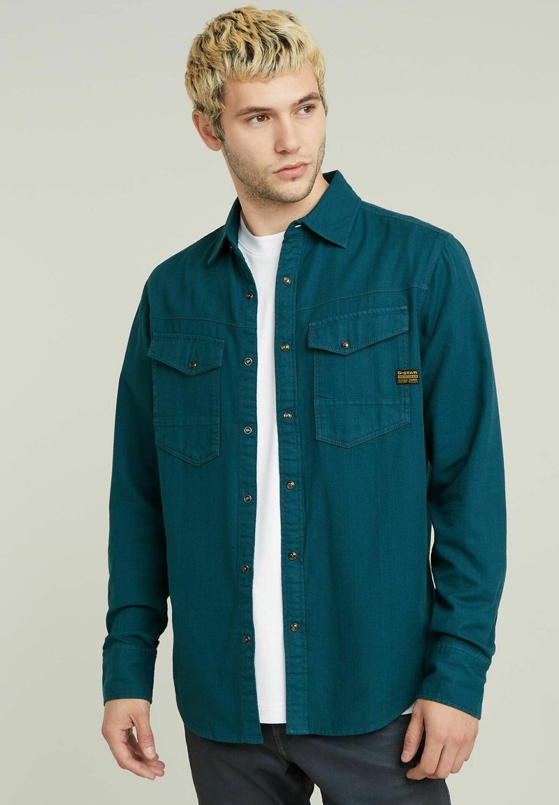 Camicia in flanella teal con maniche lunghe, apertura frontale con bottoni, due tasche sul petto e patch logo dorato. Tessuto morbido, vestibilità casual.