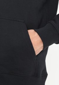 Sudadera negra con un bolsillo frontal, puños y dobladillo acanalados; fabricada en un tejido suave con una textura lisa. Primer plano de una mano en el bolsillo.
